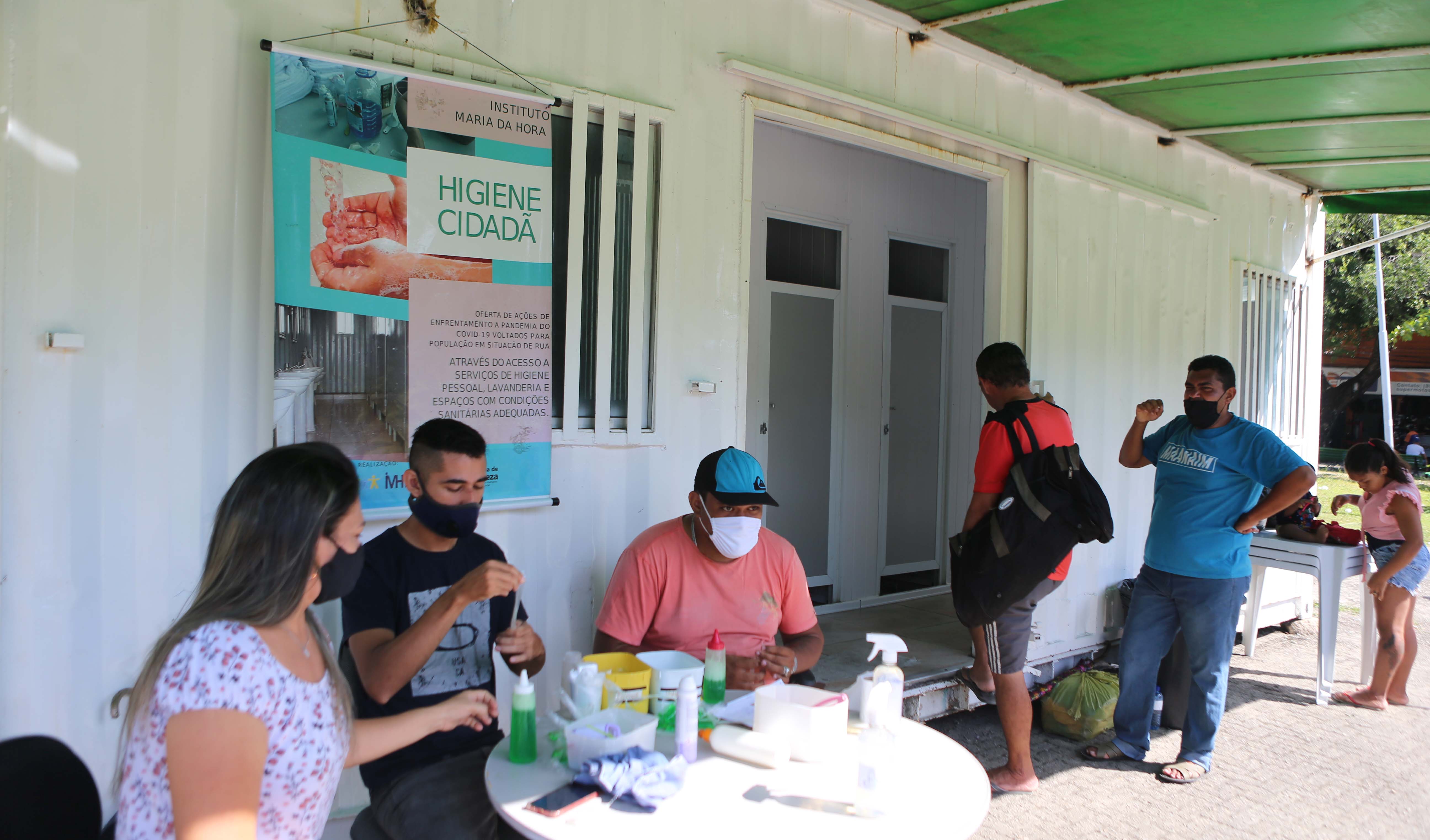 grupo de pessoas na frente do conteiner onde funciona o espaço de higiene cidadã, três funcionários da prefeitura estão sentados em uma mesa e entregando vidros de sabonete líquidos para as pessoas na fila